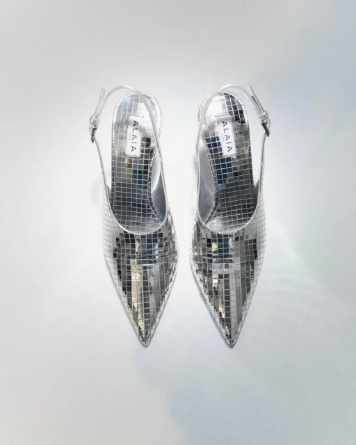 Pair of silver, pointed-toe, slingback heels, made of small reflective squares, arranged on a white background in a studio shot.
