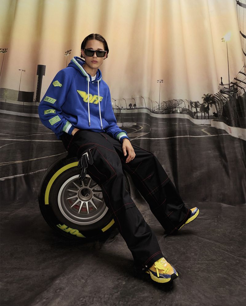 Adult posing on a tire in a blue hooded sweatshirt, black pants and trainers. Background is a race track image.