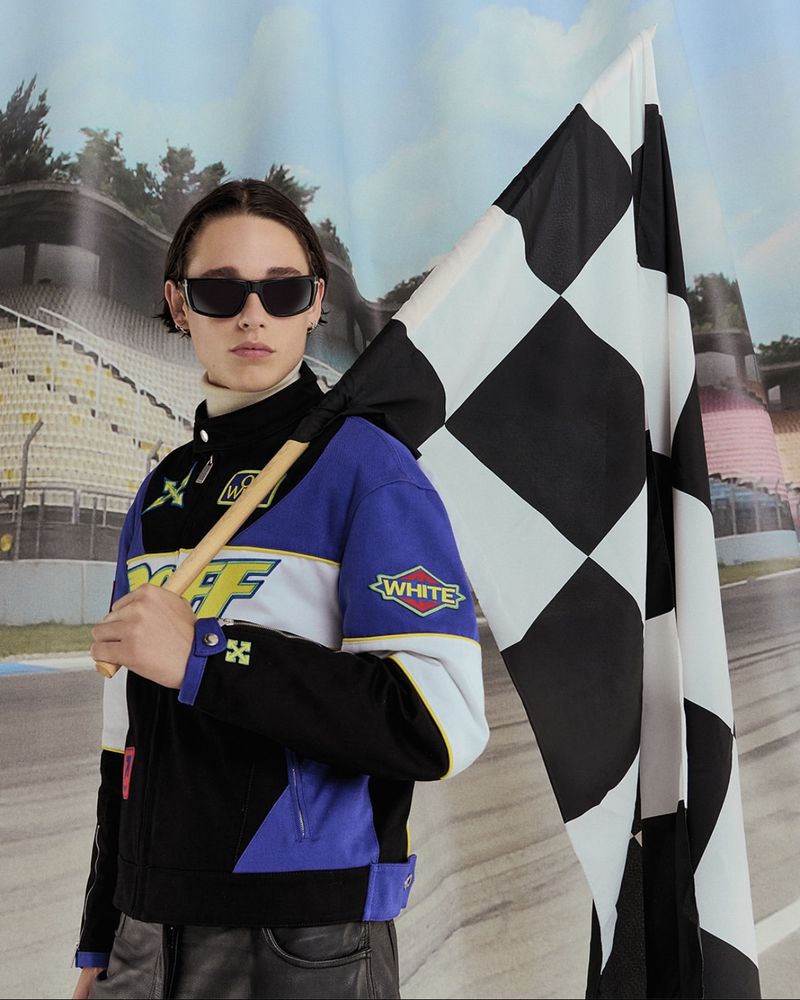 A person in a racing jacket stands holding a checkered flag against a race track backdrop, embodying a blend of fashion and sport.