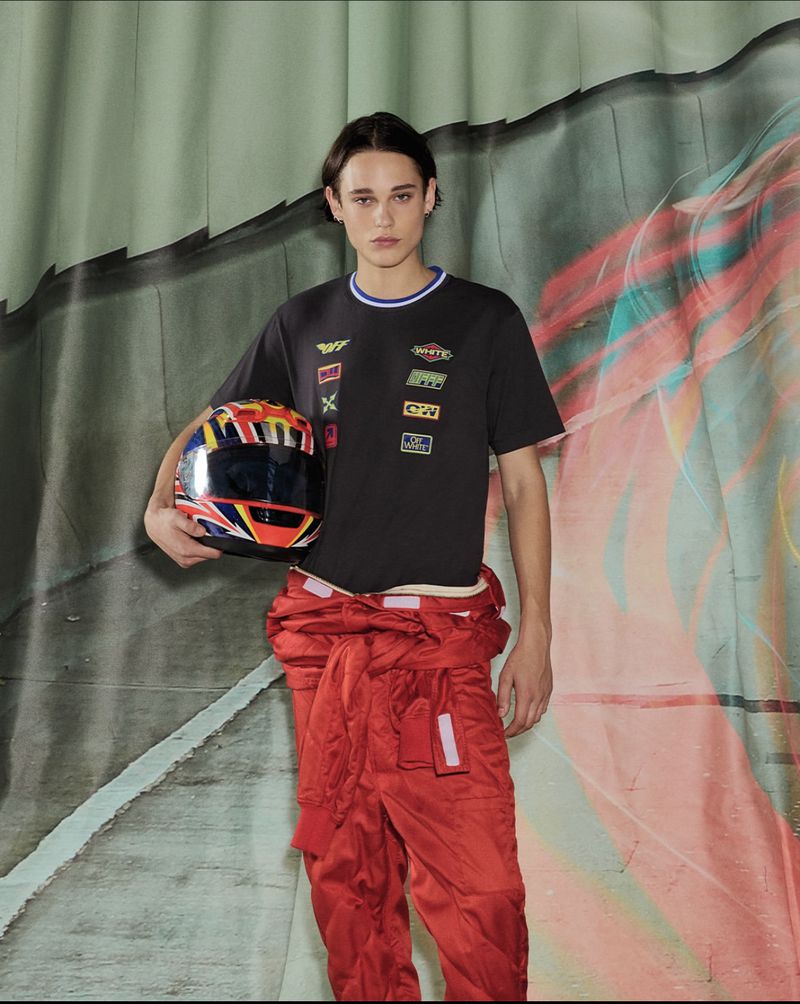Young man in a racing suit and t-shirt holding a helmet in a studio with a painted backdrop, looking directly at the camera.