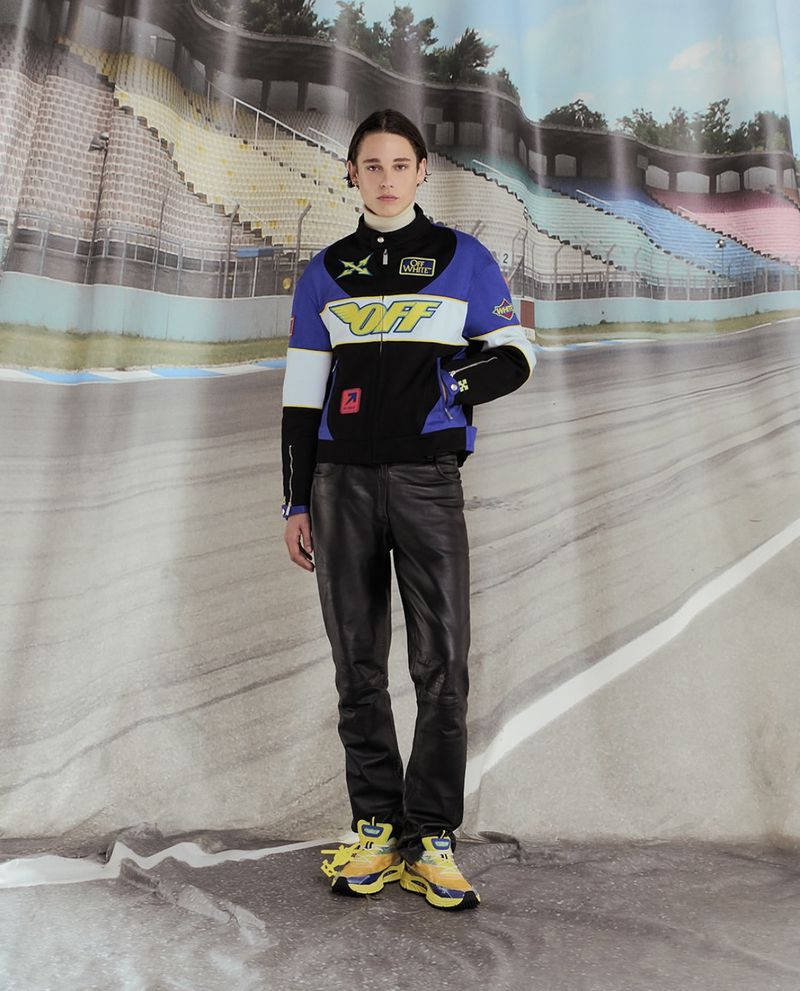 Male model in a racing-style jacket and leather pants stands against a backdrop of a racetrack, showcasing fashion and style.