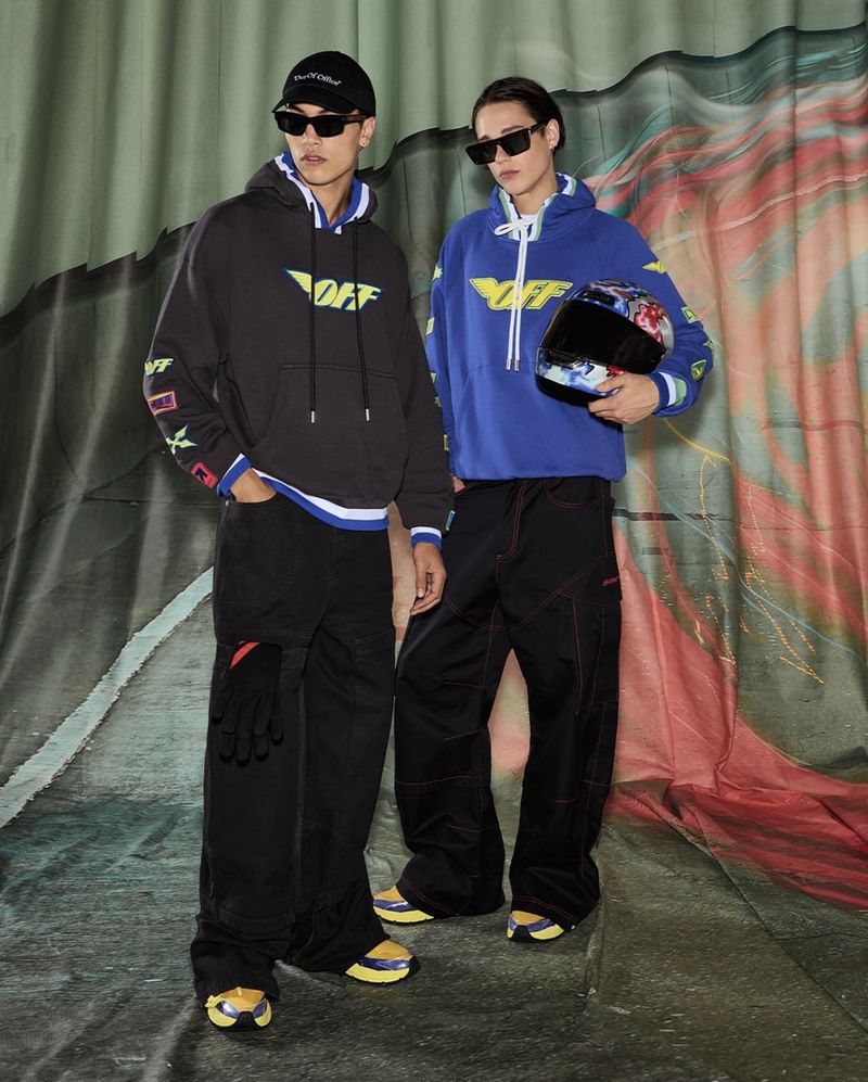 Two adult male models wear black and blue hooded sweatshirts with a stylized logo and matching black pants in a studio setting.