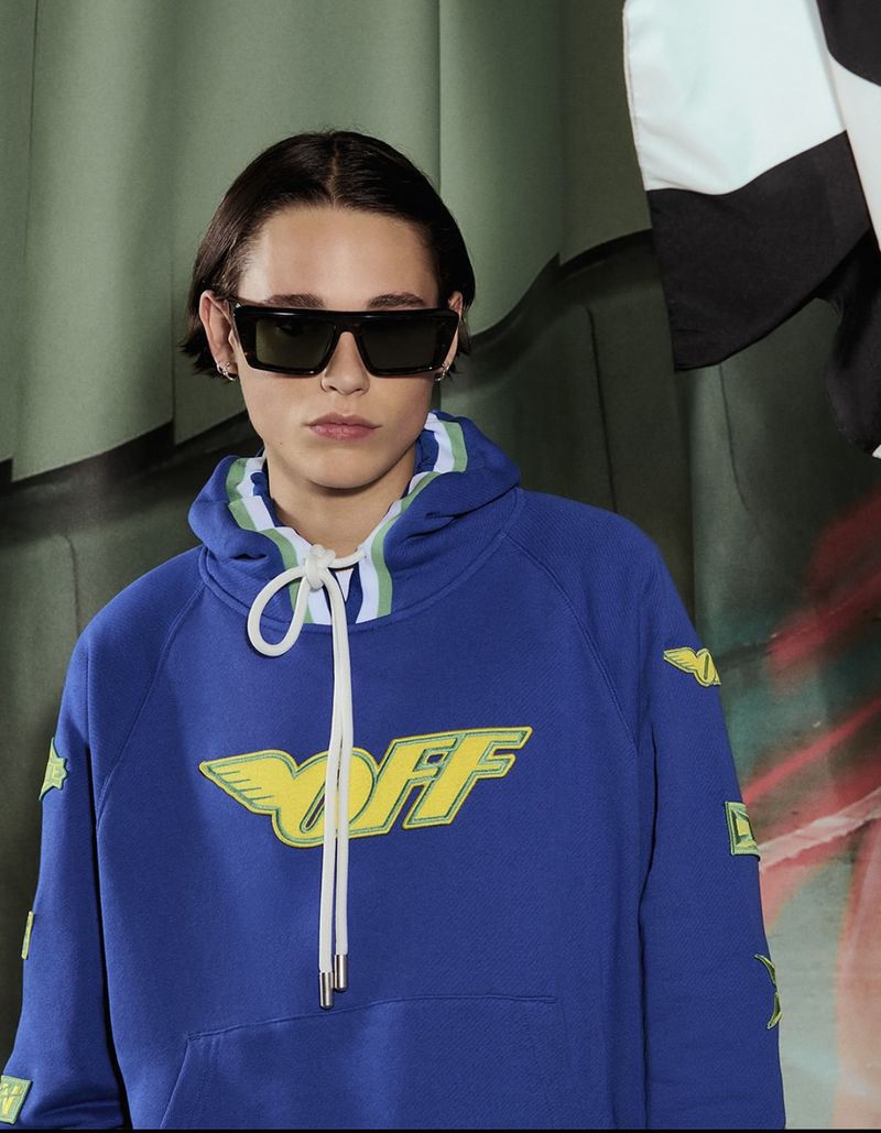 Portrait of a young adult wearing sunglasses and a blue graphic hoodie with a black and white checkered flag visible in the background.