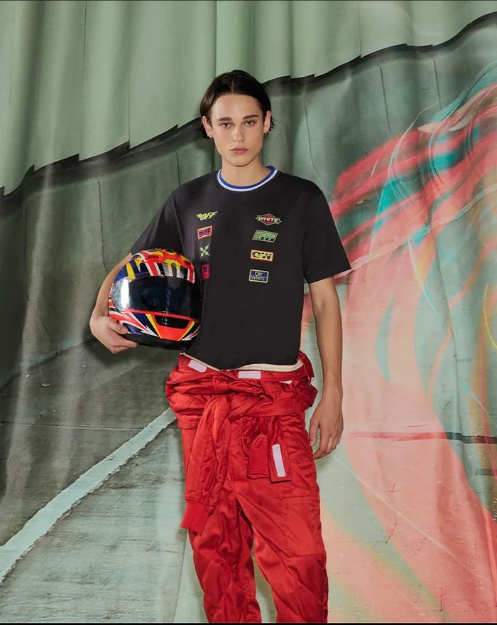 Model wearing a black t-shirt and red racing jumpsuit holding a helmet against a painted backdrop for Off White