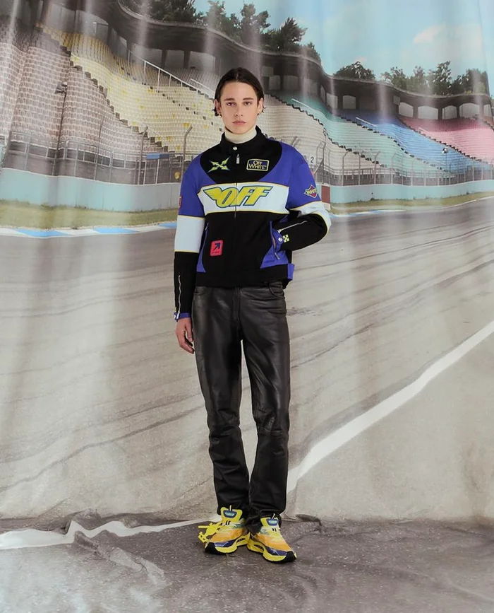 Model wearing a black, blue and white racing jacket and black trousers standing against a backdrop of a racetrack stadium