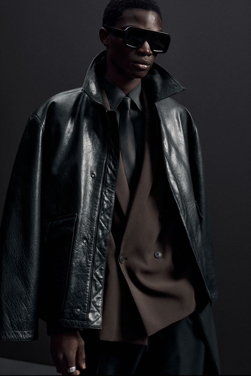 Stylish adult man wearing a black leather jacket, sunglasses, and a tie, posing in a dark studio setting for a fashion shoot.