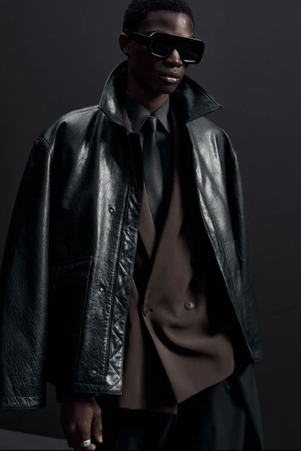Stylish adult man wearing a black leather jacket, sunglasses, and a tie, posing in a dark studio setting for a fashion shoot.