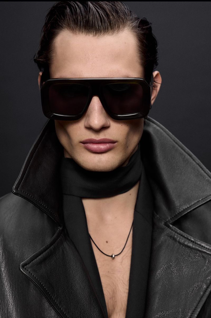 Man wearing dark sunglasses, a black leather coat, and a scarf stands against a dark backdrop in a fashion-style portrait.