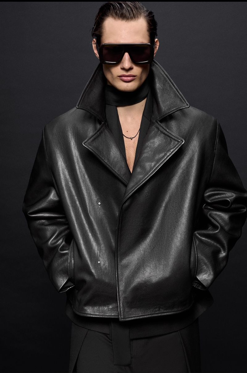 Male fashion model posing in a black leather trench coat and sunglasses in a studio with a dark background. Fashion editorial shoot.