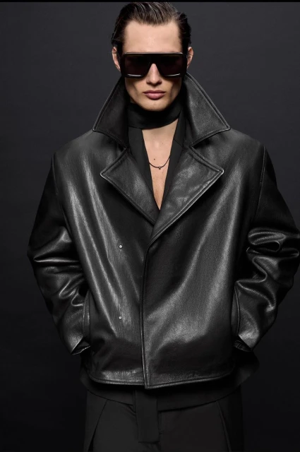 Male fashion model posing in a black leather trench coat and sunglasses in a studio with a dark background. Fashion editorial shoot.