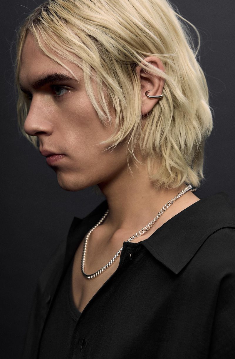 Close-up studio shot of a young adult male with light blonde hair, wearing a black shirt, silver chain necklace, and silver ear cuff.