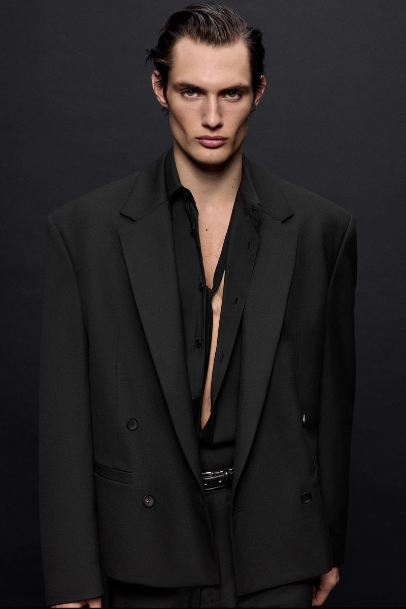 Close-up portrait of a male model wearing an oversized black blazer and unbuttoned black shirt against a dark background.