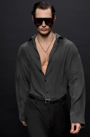 Male model wearing dark sunglasses, an unbuttoned grey shirt, and black trousers against a dark backdrop for Zara Party Winter 2024.