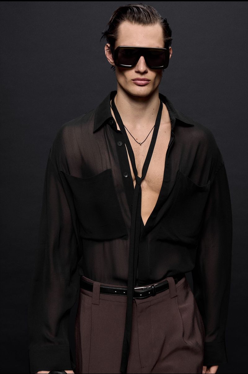 Male model wearing oversized black sunglasses and an unbuttoned sheer black shirt against a dark grey background.