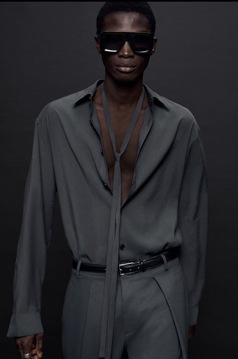 Model wearing dark sunglasses and a grey buttoned shirt with a necktie against a dark background
