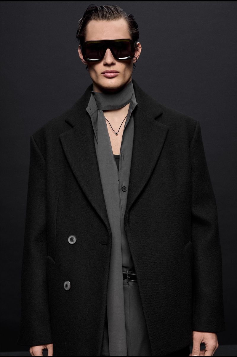 A male model poses in a black overcoat and sunglasses against a dark backdrop in a studio setting, showcasing modern fashion.