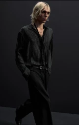 Model with blonde hair wearing a dark striped shirt and black trousers walking against a dark backdrop.