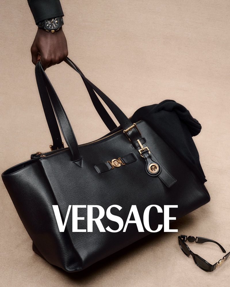 Elegant black Versace tote bag is held by an adult male hand, styled with sunglasses, exuding luxury fashion against a neutral backdrop.
