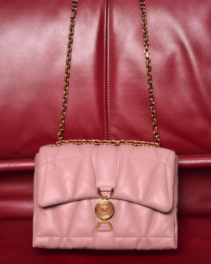 Close-up of a light pink, quilted leather shoulder bag with a gold chain strap and a gold Medusa medallion, set against a red leather background.