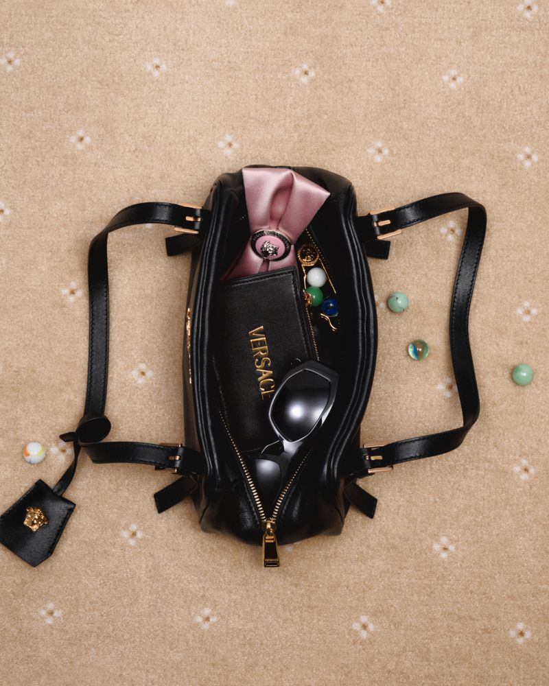 Overhead shot of a black Versace handbag with its zipper open on a beige patterned surface, filled with sunglasses and accessories.