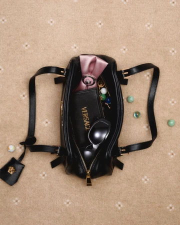 Overhead shot of a black Versace handbag with its zipper open on a beige patterned surface, filled with sunglasses and accessories.