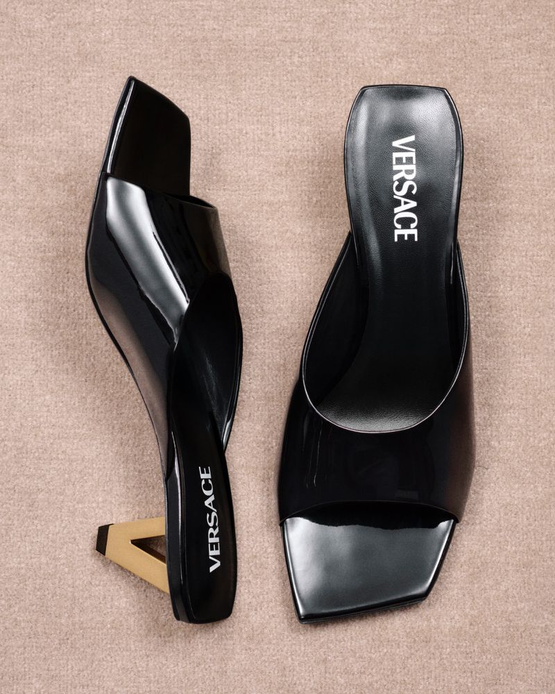 Two black Versace heeled mules with a beige backdrop. The high-fashion shoes feature a unique heel design. Fashion footwear product shot.
