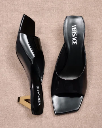 Two black Versace heeled mules with a beige backdrop. The high-fashion shoes feature a unique heel design. Fashion footwear product shot.