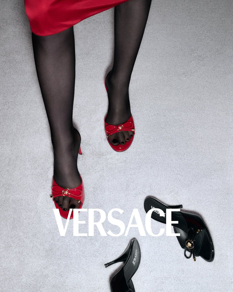 Fashion still life features a model wearing red mules and black stockings, with other black mules arranged on a gray backdrop.
