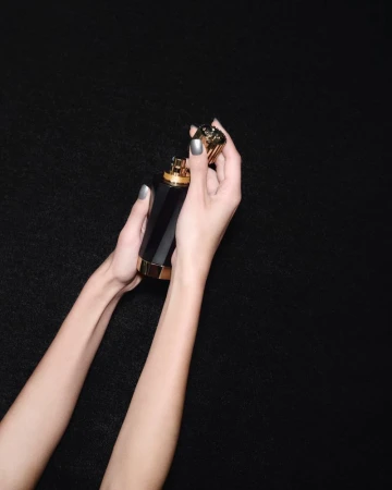 Woman's hands holding a black and gold perfume bottle against a black background, showcasing its elegant design and presentation.