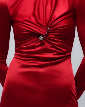 Close-up of a red satin dress with long sleeves, featuring draped fabric and a silver safety pin embellishment on the bodice.
