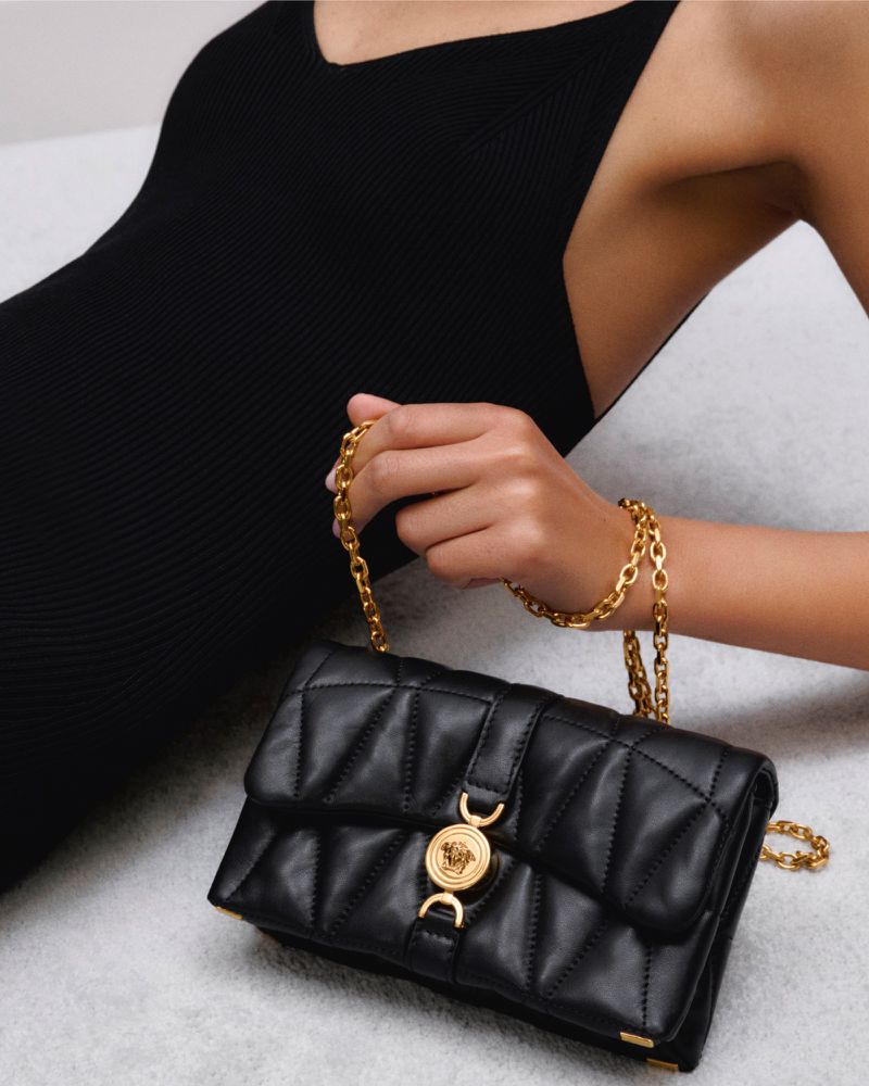 A woman in a black dress holds a black Versace quilted leather handbag with a gold chain strap and a gold Versace Medusa logo.