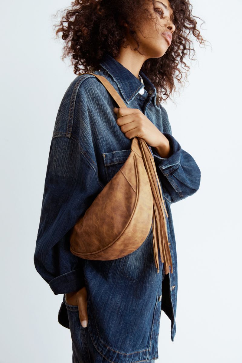 Dark-haired woman poses in a denim jacket with a tan fringe hobo bag slung across her body in a studio shot against a white backdrop.