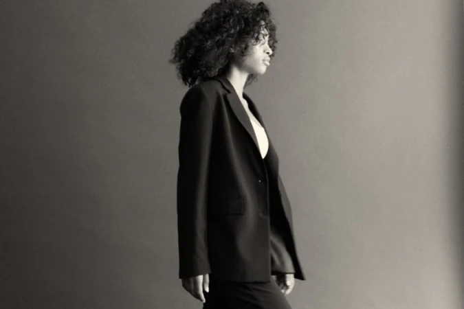 Elegant portrait of a curly-haired woman wearing a black blazer in a photo studio with a neutral backdrop and professional lighting.