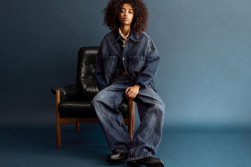 A Black female model with curly hair sits on a black leather chair, wearing a denim jacket and pants, against a dark blue backdrop.