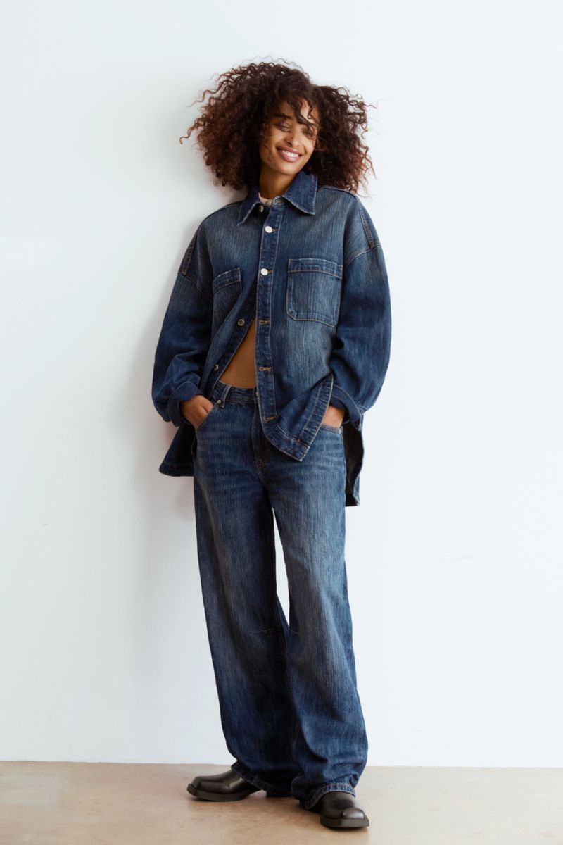 A young adult woman with curly hair smiles in an oversized denim shirt and jeans against a white backdrop in a studio portrait.