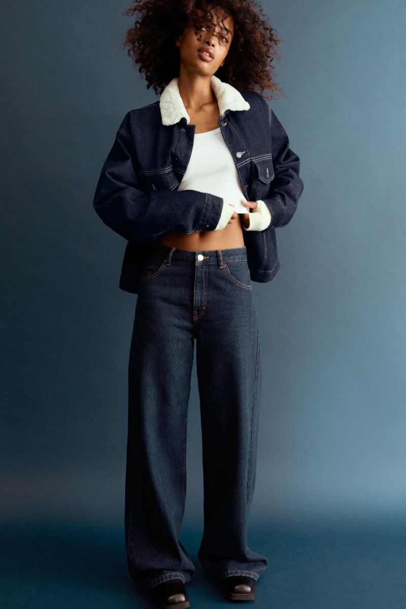 Fashionable woman poses indoors in a denim jacket with a white collar, jeans, and a white top in a studio setting.