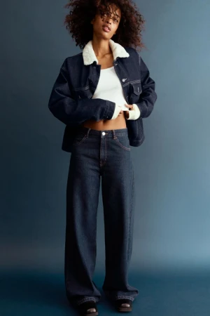 Fashionable woman poses indoors in a denim jacket with a white collar, jeans, and a white top in a studio setting.