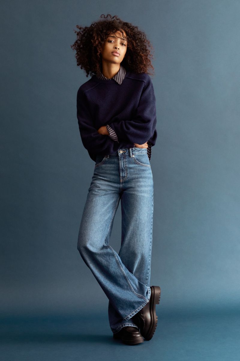Full-length studio shot of a stylish, young woman with curly hair wearing a dark blue sweater, jeans, and black platform shoes.