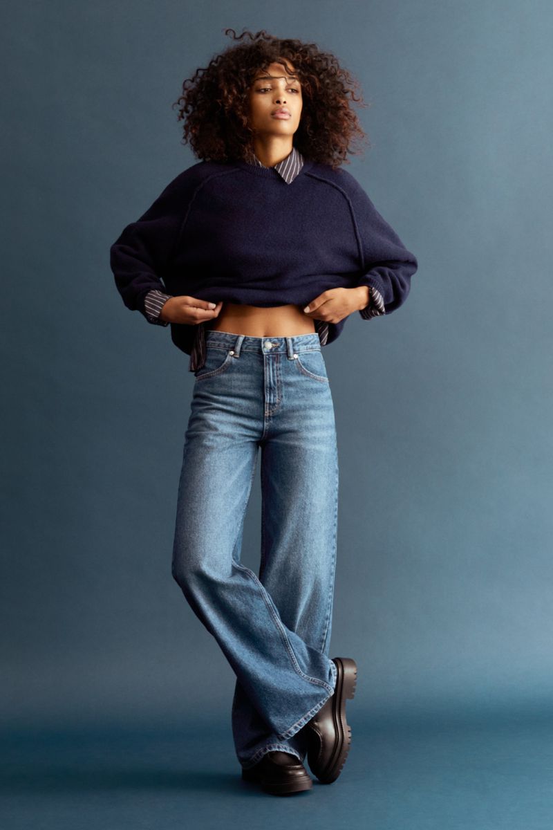 A woman poses in a navy sweater, blue jeans, and black shoes against a muted blue background in a full studio shot.