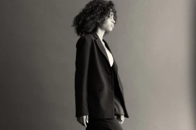 Model with dark curly hair wearing a black blazer and trousers against a grey seamless background