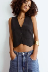 Model with curly brown hair wearing a dark pinstripe vest and denim jeans against a plain white background