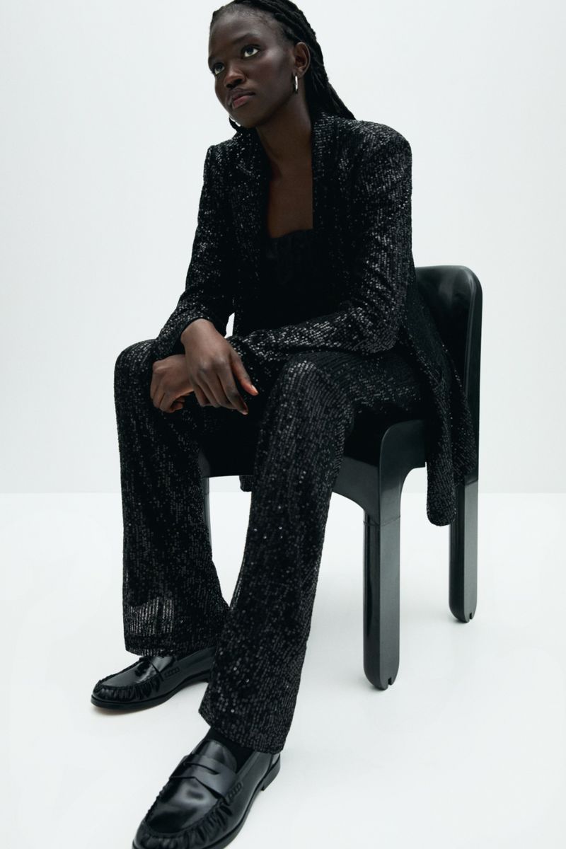 A Black woman sits on a chair wearing a sequined blazer and pants with loafers against a white background in a studio setting.