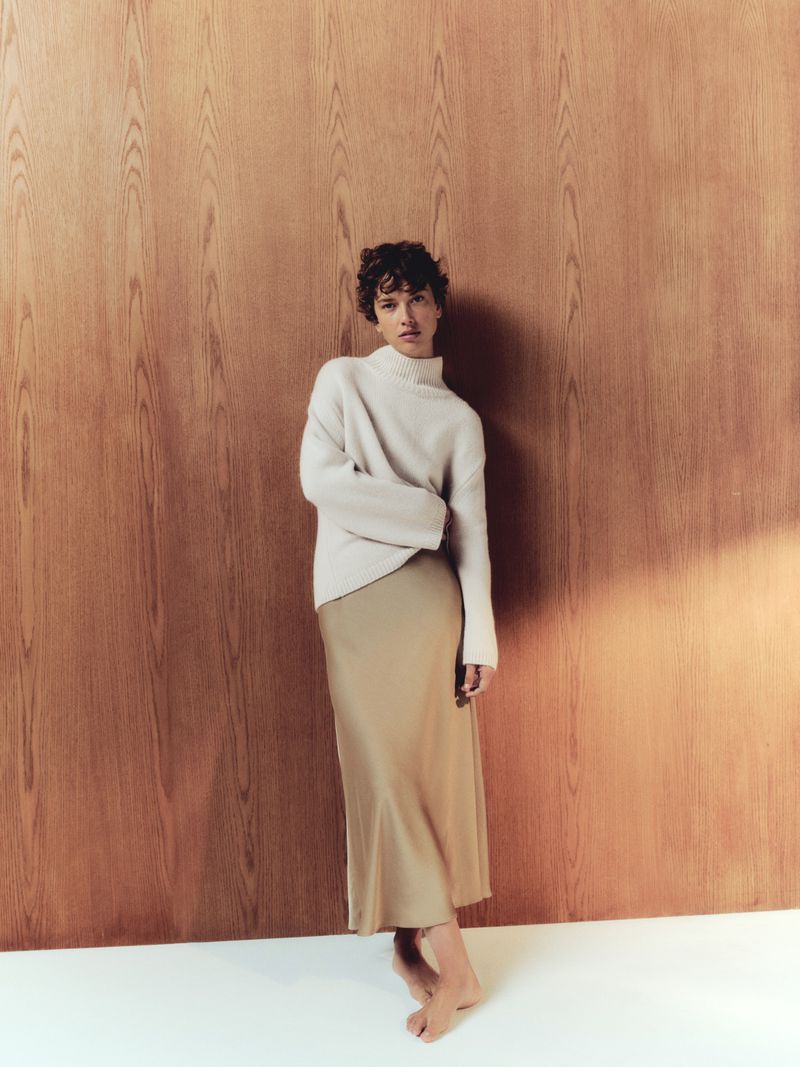 A woman with short brown hair is standing barefoot, wearing a beige sweater and silk skirt, posed in front of a wooden wall.