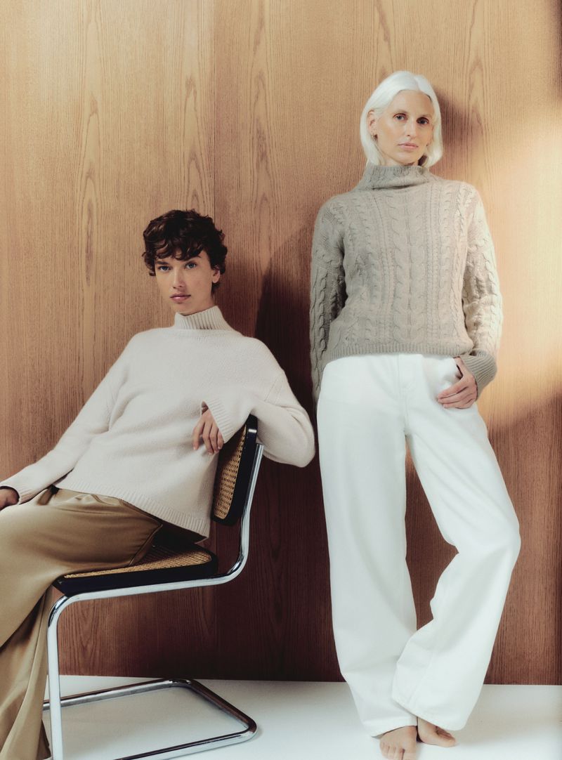 Two women in knit turtlenecks and trousers pose against a wood-paneled wall. One woman sits on a modern chair; the other stands.