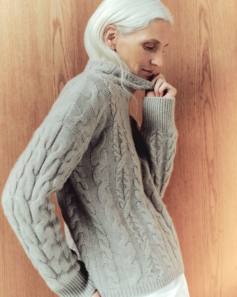 Elegant elderly woman with white hair in a gray cable knit turtleneck sweater posing in front of a wood panel wall in a fashion shot.