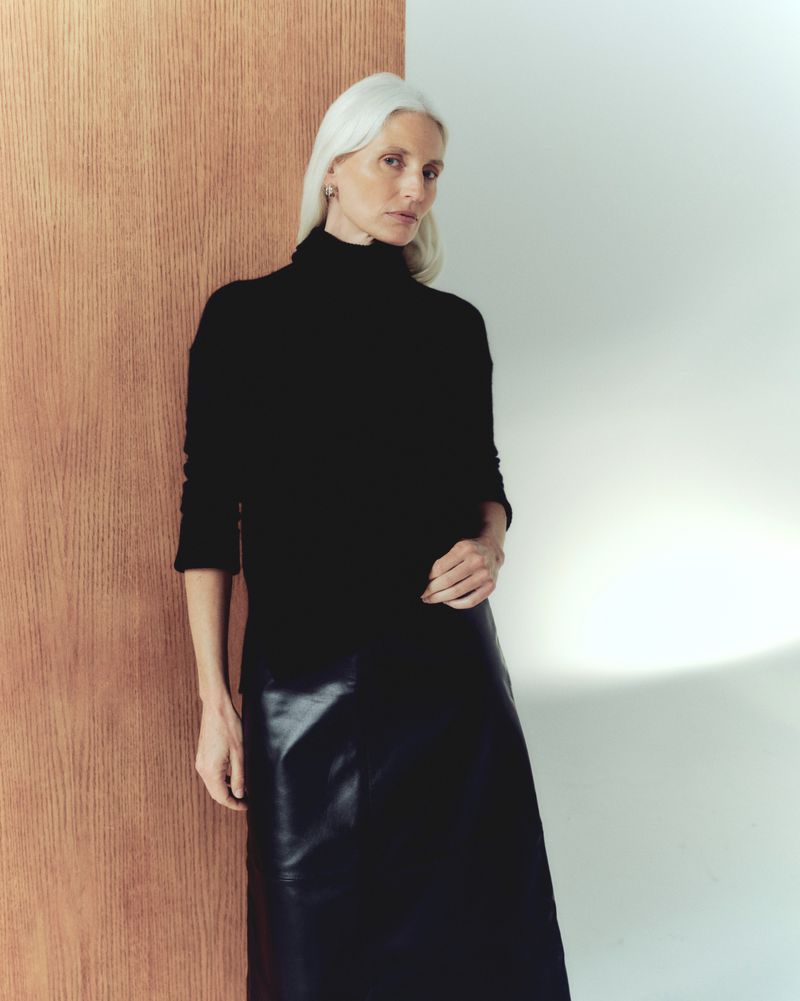 Elegant woman with platinum hair poses in a black turtleneck and shiny black skirt against a neutral background with natural light.