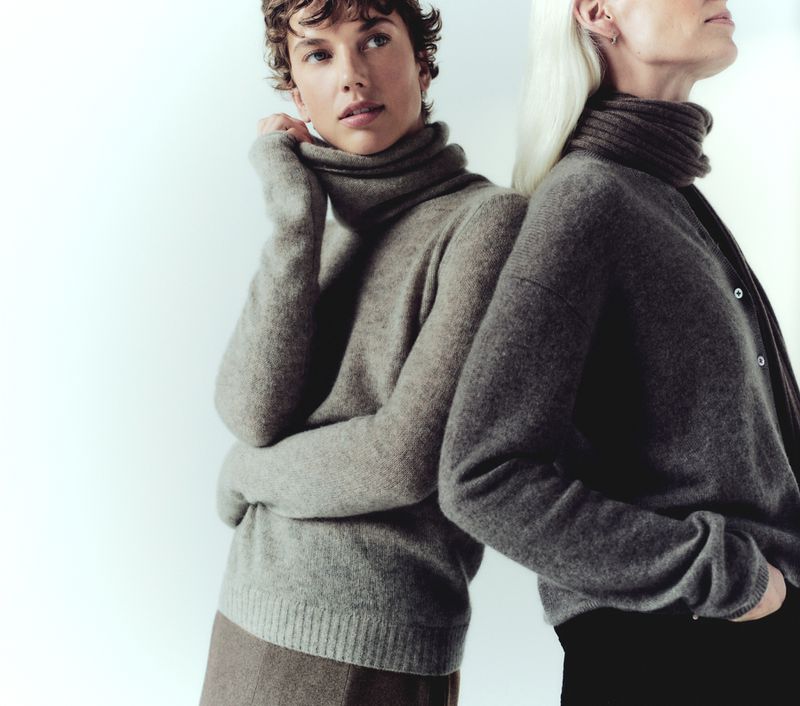 Two fashion models pose back-to-back in a studio wearing elegant knitwear in neutral tones. Focus on textures and fabric details.