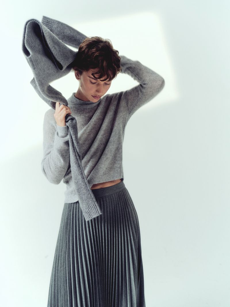 A young adult woman in a gray sweater and pleated skirt puts on a gray scarf in a bright, minimalist studio, fashion editorial style.