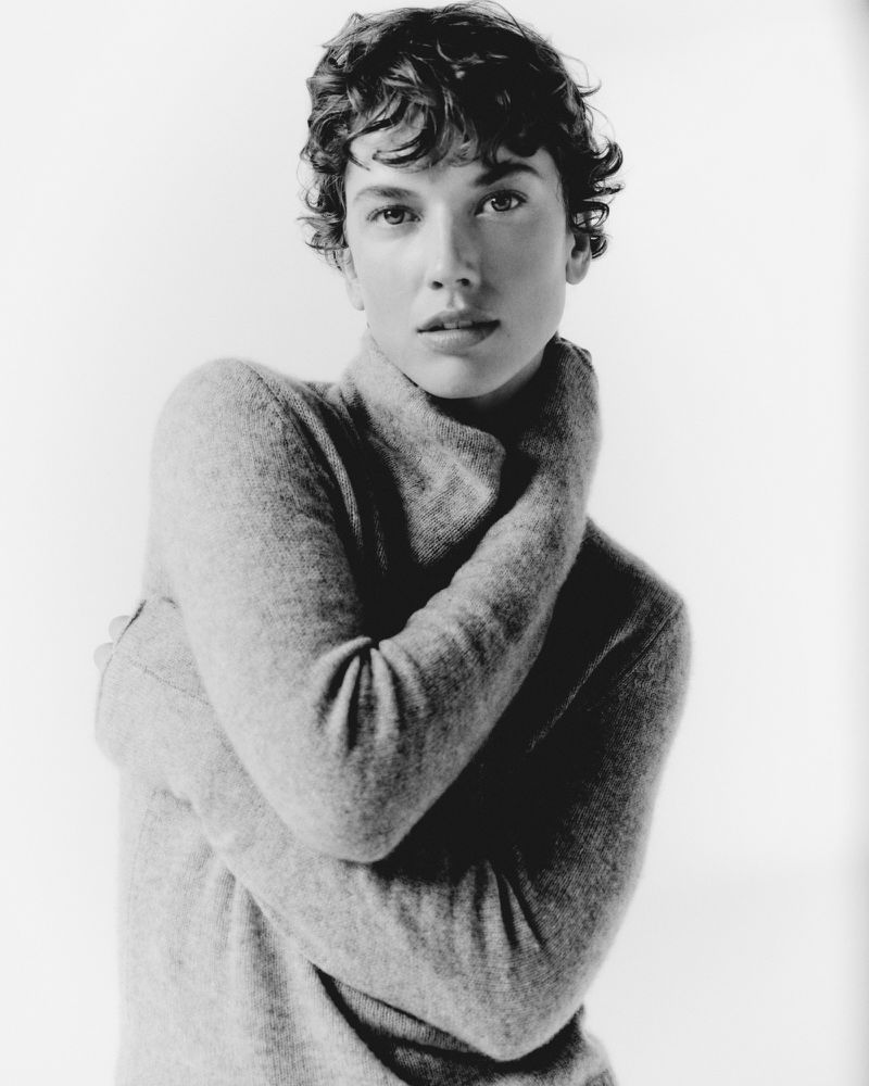 Model with short curly hair wearing a grey cashmere turtleneck sweater against a white background