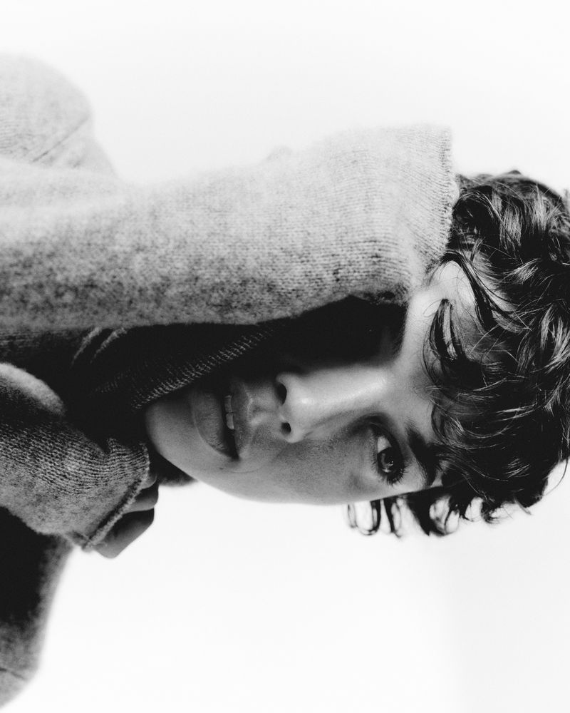 Close-up portrait in black and white shows a young adult with curly hair, eyes open, wearing a sweater, against a white background.